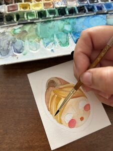 mano che colora l'acquerello da inserire nel ciondolo di carta rappresentante un viso di una bambola