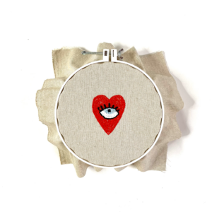 pupilla azzurra dell'occhio dentro cuore rosso con filo rosso infilato in punch needle