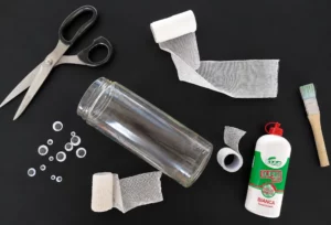 Materiali per realizzare una lanterna mummia con un barattolo di vetro, forbice, occhi in plastica, colla, pennelli e garze