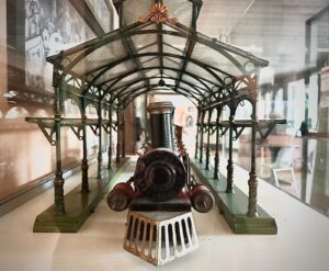 Trenino vintage visto frontalmente sotto una pensilina ferroviaria verde in ferro e vetro